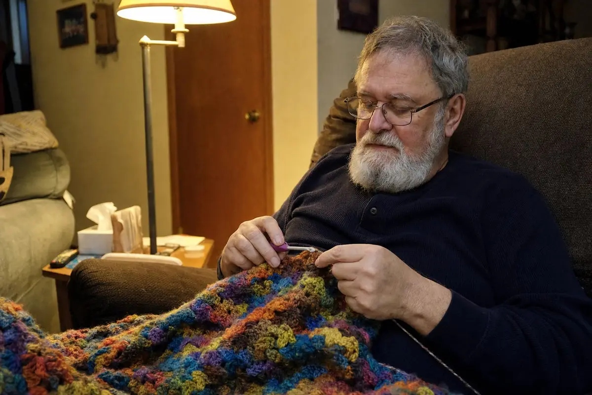 An older man crochets a blanket with a thick-handled hook in his armchair