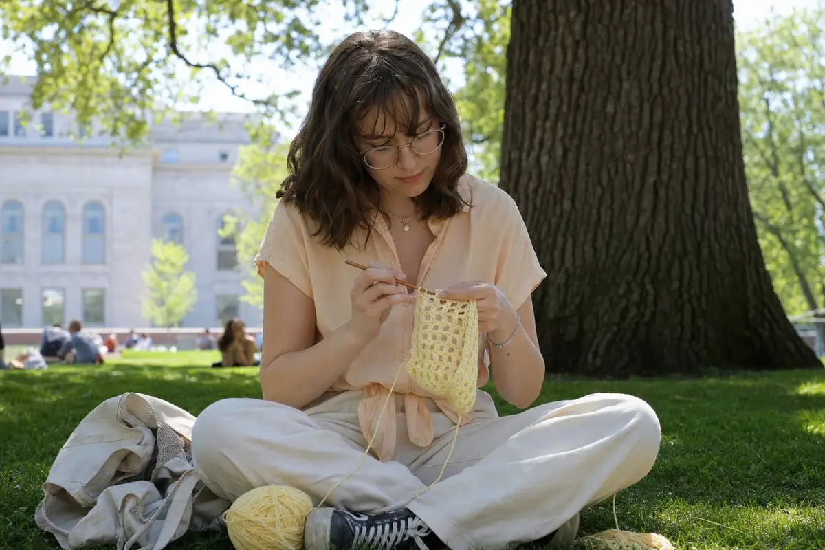 A woman works tall double crochet stitches on a butter-yellow swatch on a campus lawn