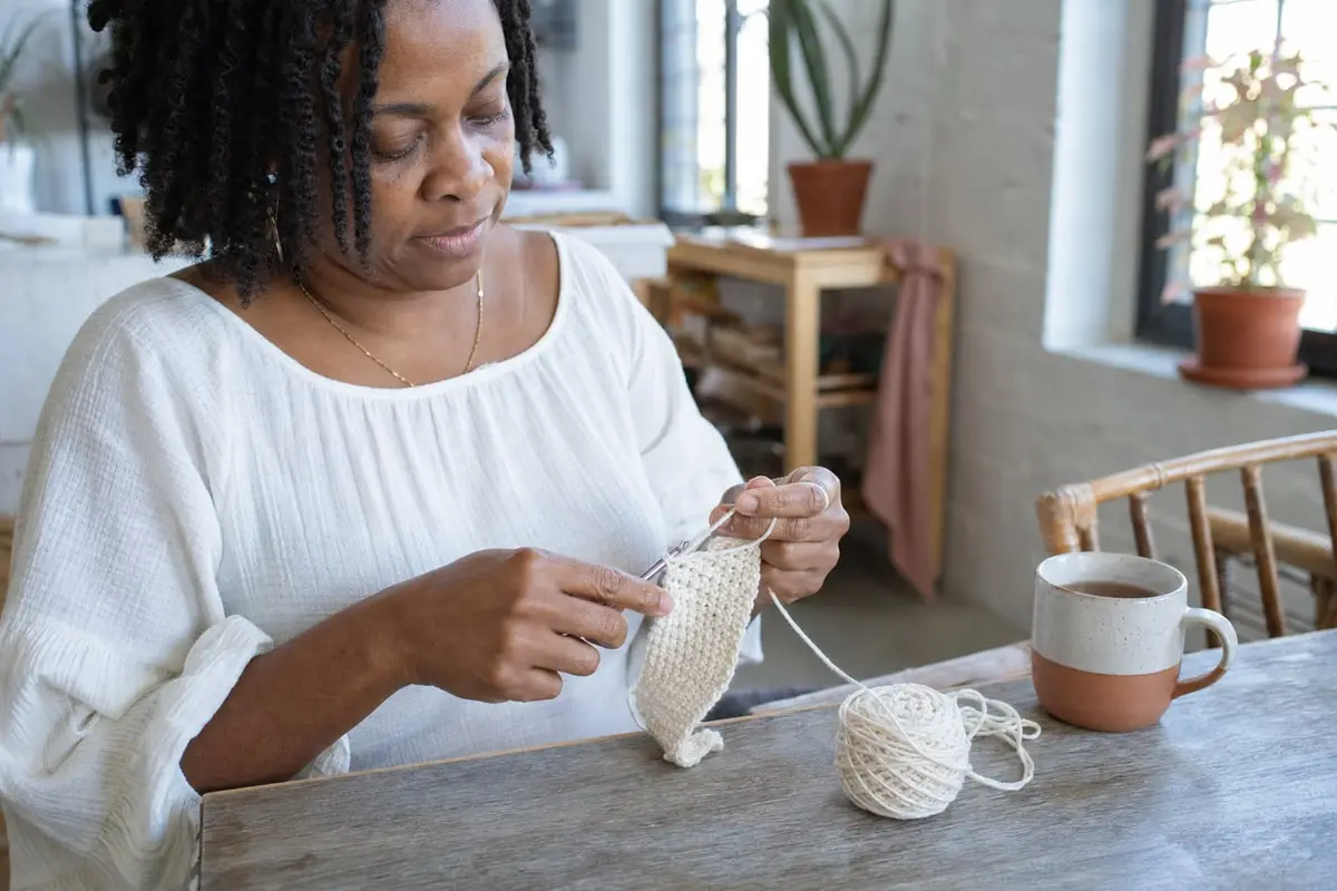 Hands work a row of single crochet on a small cream swatch at a kitchen table